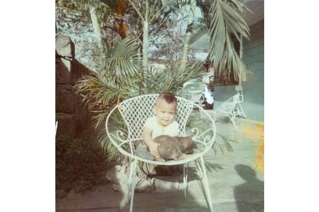 Male toddler in a large chair, outside his house, with a puppy in his lap and palm trees behind him.